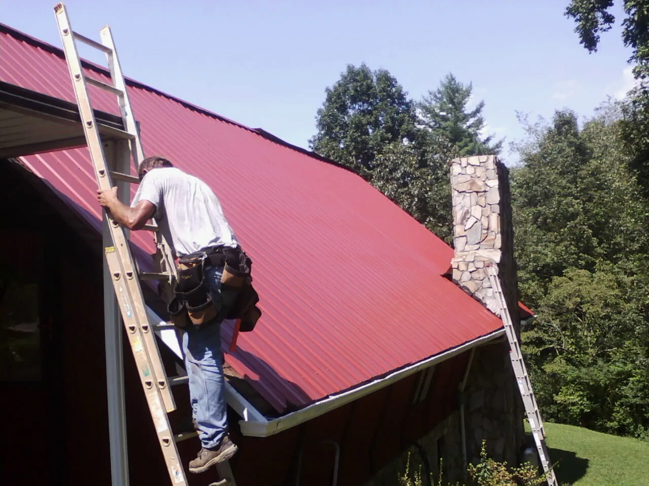 Roofer on a ladder beside a red metal roof and stone chimney — Soffit & Fascia Repair by Roofing Brothers in Ruskin